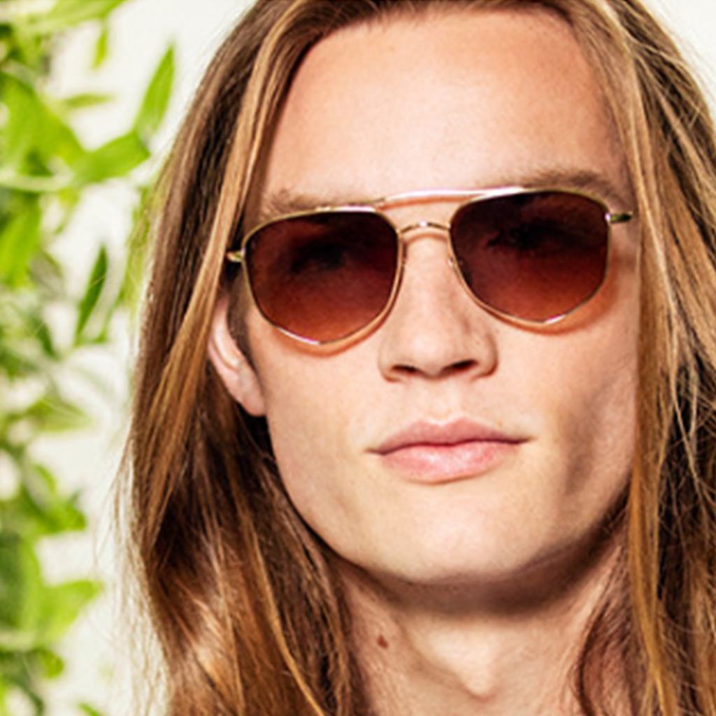 Homme aux cheveux longs portant des lunettes de soleil à monture dorée avec des verres dégradés marrons, avec un fond de feuilles vertes floues